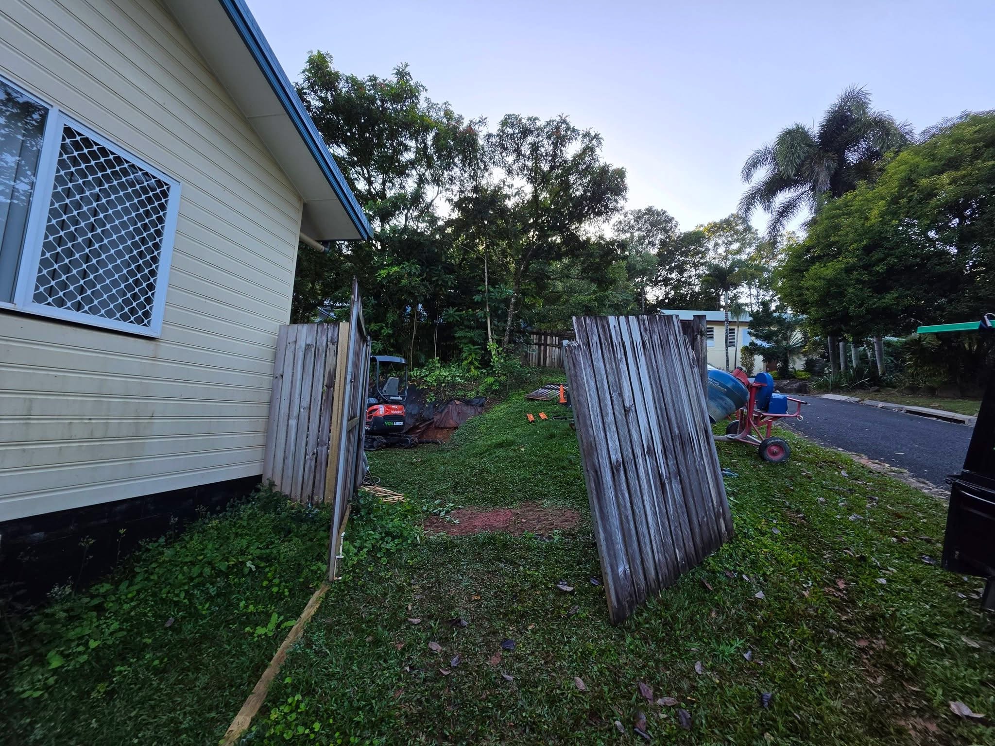 Old timber fence panels lifted out and leaned up, with a mini excavator and cement mixer on site