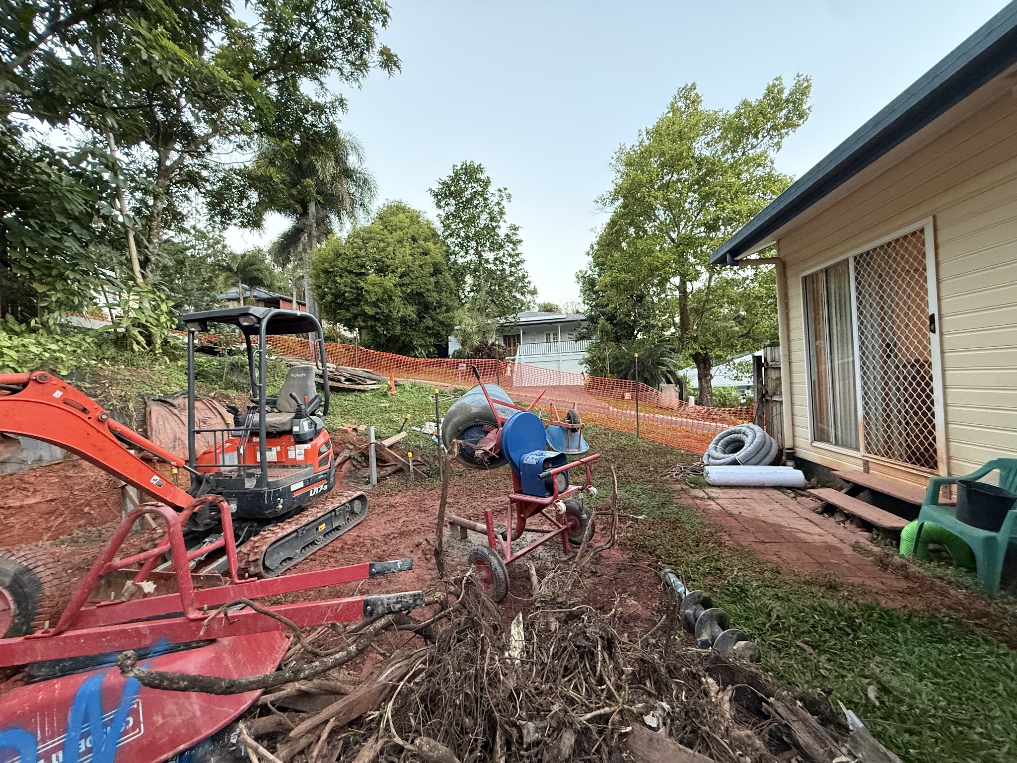 A Kubota mini excavator and a cement mixer on a job site with orange safety mesh and stump grindings